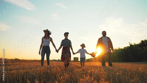 Family walking on wheat field at sunset. Happy parents children holding hands talking together in summer vacations. Farmers having fun together on farm. Farming lifestyle, recreation, leisure concept. photo