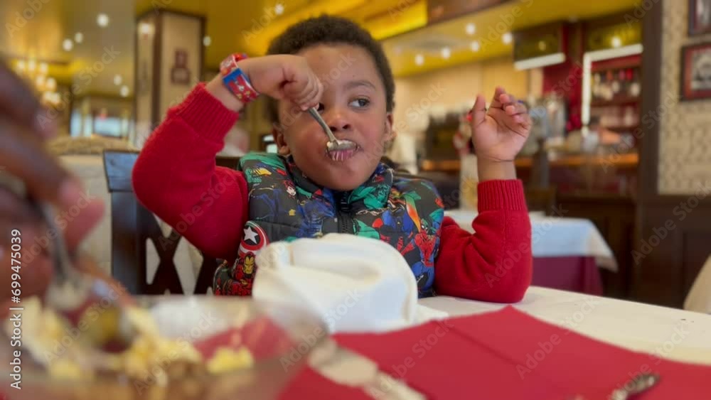 3yearold black kid eating a tiramisu dessert in an elegant italiant restaurant seated next to