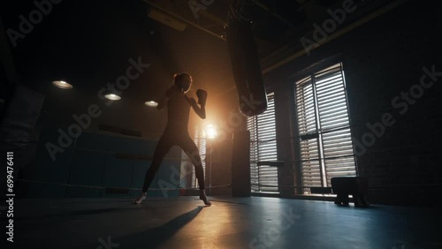 Woman boxer practicing punches using punching bag in gym on boxing ring, bottom view. Young focused girl professional fighter has hard training, full height. Workout, box, sport fighting concept. photo
