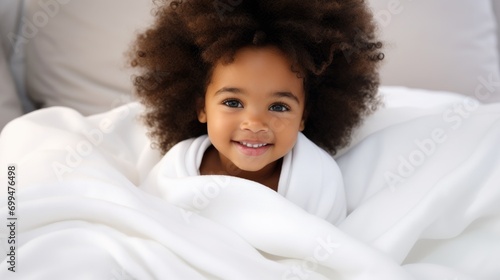 A very cute little black african baby kid with afro hair wrapped in soft white blanket on a bed smiling. image perfect for ads. big beautiful eyes and tiny nose