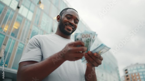Man counts dollars banknotes on street. Contented smiling african american guy enjoying earned cash money in city business center, low angle view. Finances, wages concept. Entrepreneur, businessman. photo