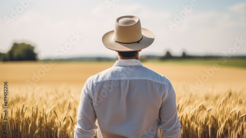 A adult white american farmer man standing on a wheat grass field. wearing a hat. photo taken from behind his back. agricultural land owner. blurry field and a mansion background © Zainab