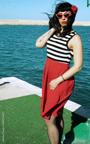 Port harbor pin-up: Red skirt, stripes, retro glasses. Red heels, intriguing secrets beneath: garter belt and sheer stockings. Revisited vintage elegance.