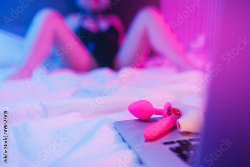 Beautiful woman lies in bed holding clitoral vibrator and watching porn on laptop, neon color. Women has Internet sex chat online photo
