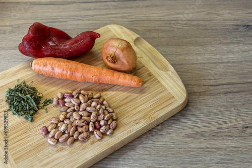 Top view of a handful of kidney beans, dried spearmint, red pepper, carrot and onion lied down on a wooden cutting board.