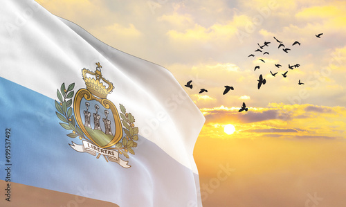Waving flag of San Marino against the background of a sunset or sunrise. San Marino flag for Independence Day. The symbol of the state on wavy fabric.