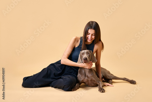 Loving girl with dog having fun. Portrait of young beautiful woman playing with her adorable Weimaraner against beige background. Concept of animal, pet lover, friendship, domestic life, companionship