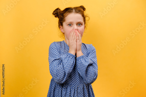 Omg. Portrait of surprised preteen girl child opening mouth and touching face, looking at camera in shock, posing isolated over plain yellow color background wall in studio. People emotion concept photo