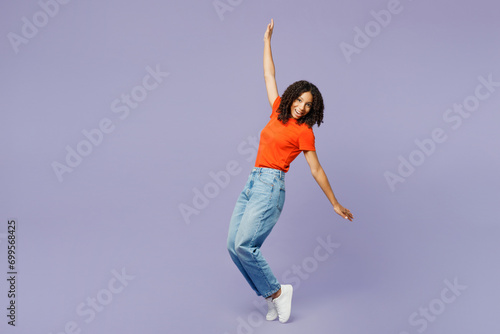 Full body little kid teen girl of African American ethnicity wear orange t-shirt stand on toes with outstretched hands lean back isolated on plain pastel purple background Childhood lifestyle concept photo