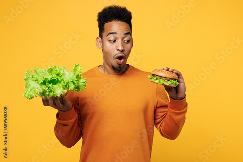 Young shocked man wear orange sweatshirt casual clothes hold lettuce salad burger choose what to eat isolated on plain yellow background. Proper nutrition healthy fast food unhealthy choice concept. photo