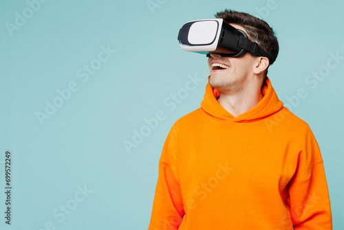Young smiling happy man he wears orange hoody casual clothes watching in vr headset pc gadget look aside isolated on plain pastel light blue cyan color background studio portrait. Lifestyle concept. photo