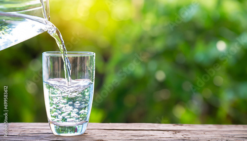 Water from jug pouring into glass on wooden table outdoors. Drink water pouring in to glass over sunlight and natural green background. Photo select focus with copy space.