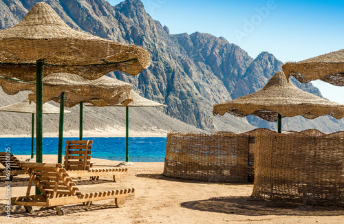 beach umbrellas and wooden lounge chairs on the sand of the beach against the backdrop of the sea and high rocky mountains in Egypt