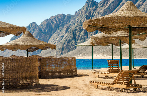 beach umbrellas and wooden lounge chairs on the sand of the beach against the backdrop of the sea and high rocky mountains in Egypt