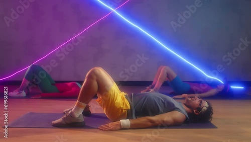 Pan shot of sportspeople in retro activewear performing glute bridges while exercising on floor at spacious neon studio photo