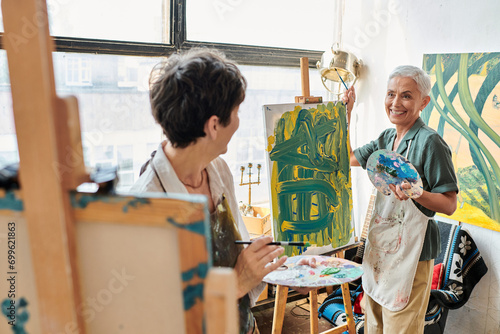joyful mature women looking at each other near easels in art studio, artistic creative hobby