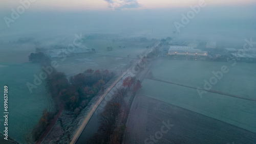 The image presents a captivating early morning scene where a dense mist blankets a rural landscape at dawn. A straight road cuts through the scene, dividing the frost-covered fields that display