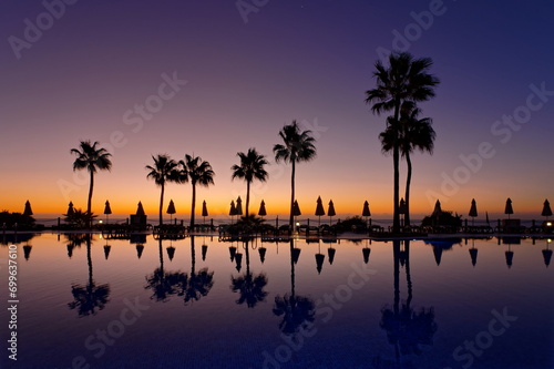Morgendämmerung am Pool mit Spiegelung auf dem Wasser, Palmen und Sonnenschirme im Hintergrund