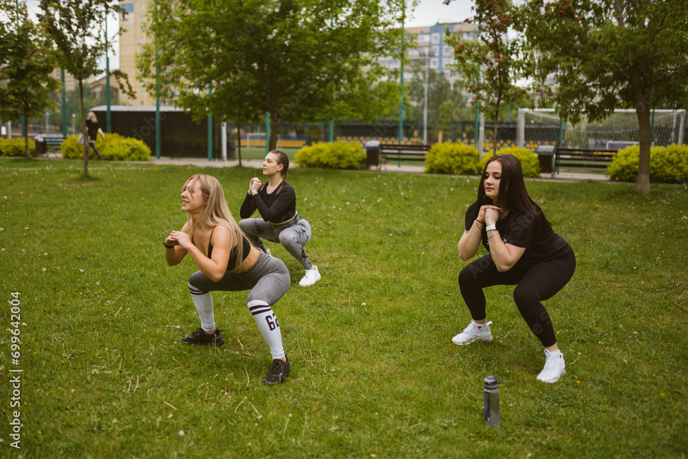 Fototapeta premium Happy female athletes do squat exercises in morning square. Young women do cardio training to refine figure. Healthy habits concept
