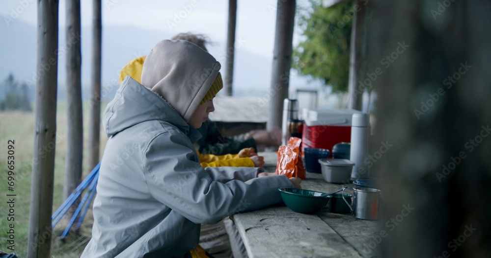 Young boy sits at the table with family and holds hiking food in pouch. Group of tourists rest in gazebo after long walking trek. Hikers during expedition to mountains in autumn. Outdoor recreation. Young boy sits at the table with family and holds hiking food in pouch. Group of tourists rest in gazebo after long walking trek. Hikers during expedition to mountains in autumn. Outdoor recreation.