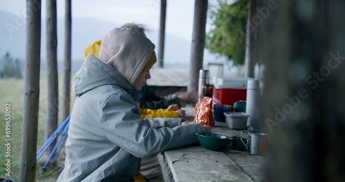 Young boy sits at the table with family and holds hiking food in pouch. Group of tourists rest in gazebo after long walking trek. Hikers during expedition to mountains in autumn. Outdoor recreation.