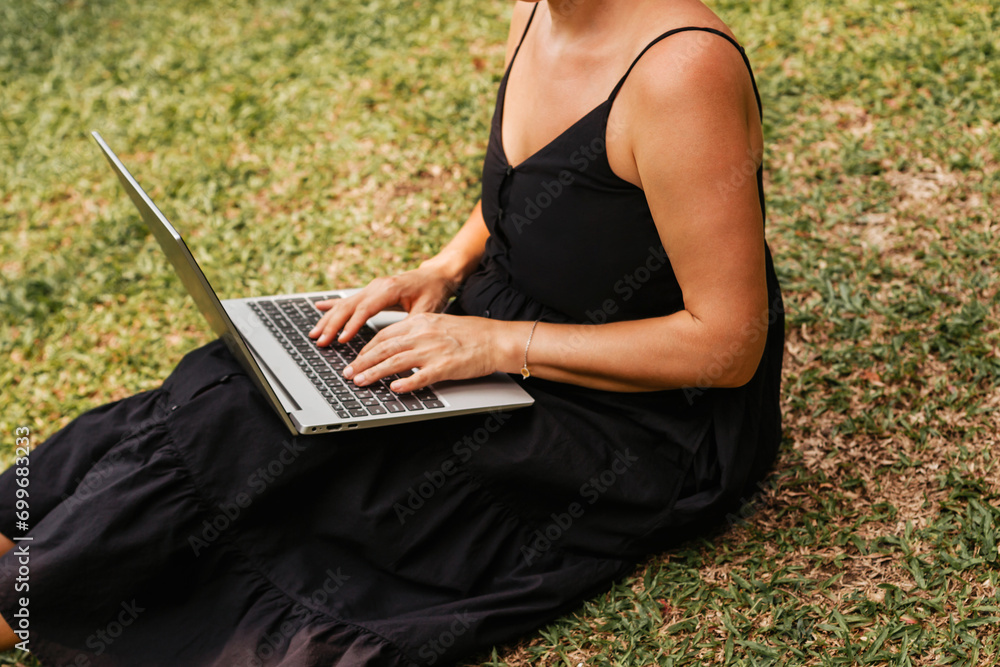 Naklejka premium Young woman typing on grey laptop sitting in summer in park