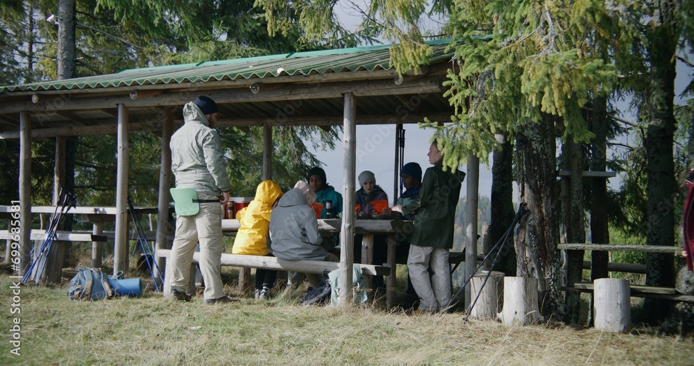 Diverse hiking buddies talk and prepare hiking food in pouches for dinner. Group of tourists rest in gazebo after walking trek in the forest. Hikers during trip or expedition to mountains on vacation.