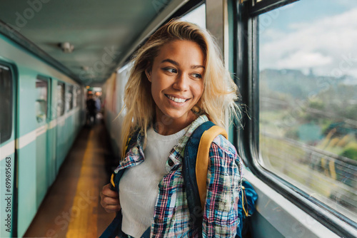 Junge lächelnde Frau mit blonden Haaren und Rucksack auf Reisen in einem Zug am Fenster photo