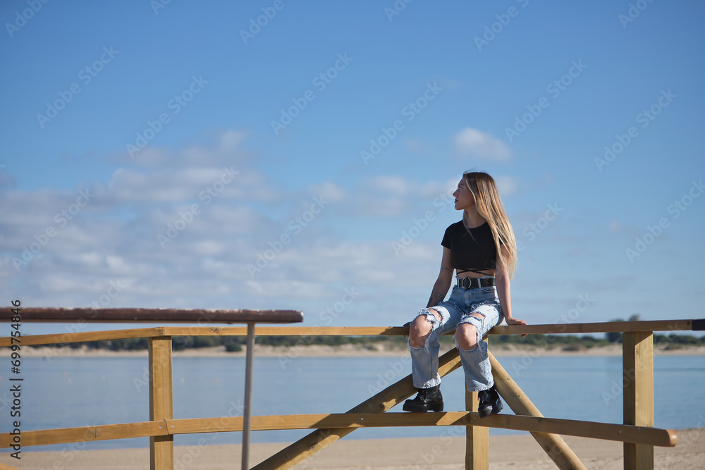 Young, pretty, blonde woman, wearing a black top, ripped jeans and tattoos, sitting on a railing, looking at infinity with the sea in the background. Concept of peace, tranquility, relaxation.