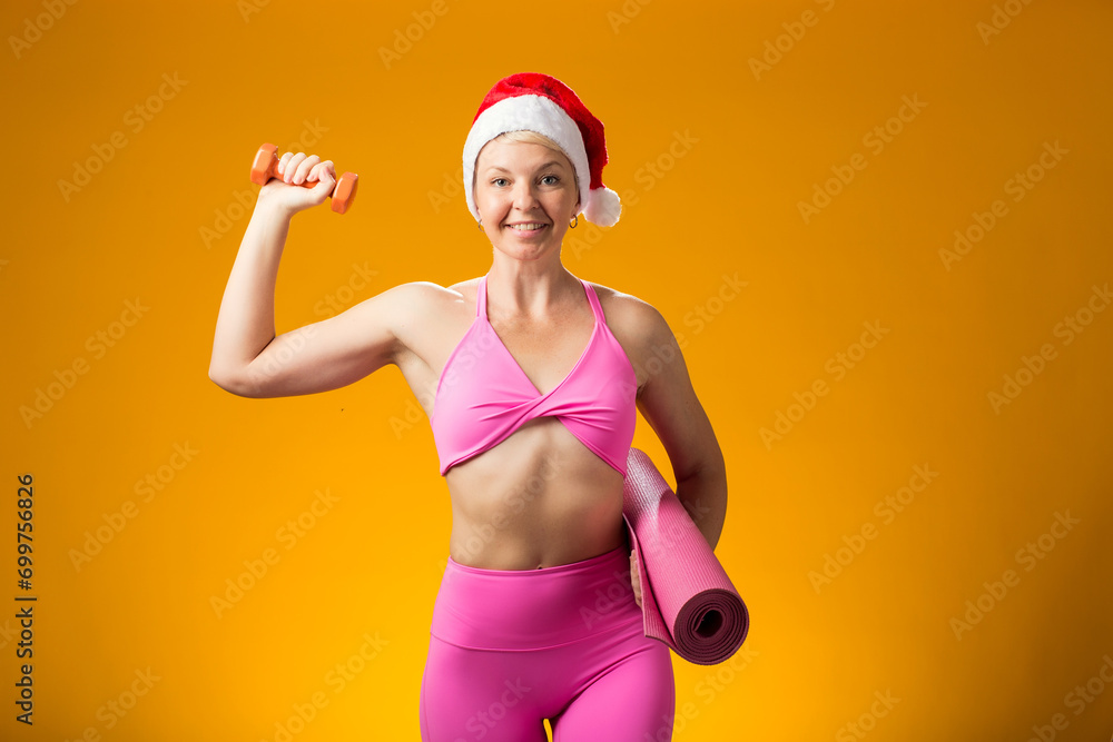 Woman in Christmas hat holding dumbbell and yoga mat isolated on yellow background. Holiday and fitness concept Woman in Christmas hat holding dumbbell and yoga mat isolated on yellow background. Holiday and fitness concept