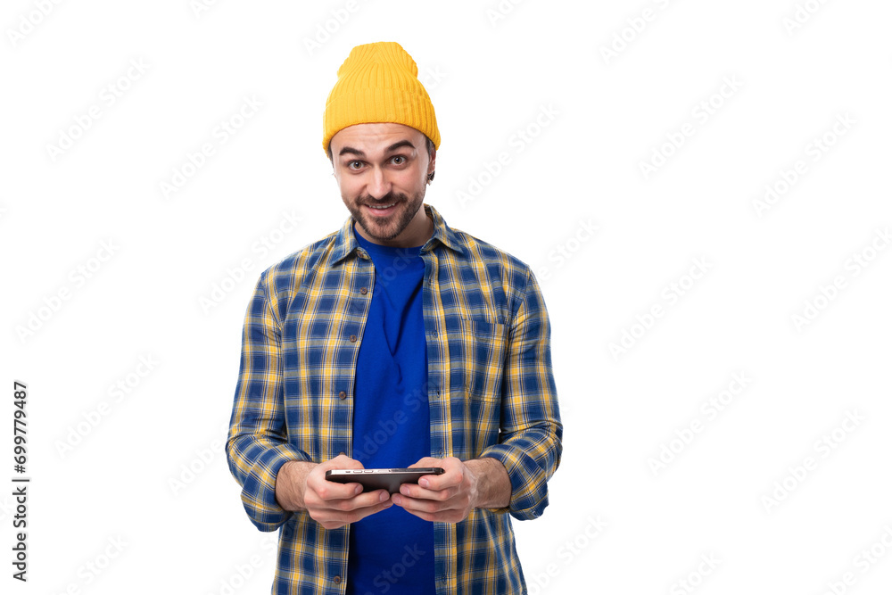 well-groomed young European brunette man with a beard and mustache dressed in a yellow hat and blue shirt uses social networks in a smartphone well-groomed young European brunette man with a beard and mustache dressed in a yellow hat and blue shirt uses social networks in a smartphone