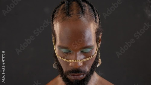 Black gay stands isolated on black background stares into camera with serious expression on face. Close-up portrait of handsome adult transgender with blue eyeshadows and golden accessory on face photo