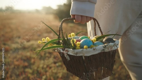 Painted Easter Eggs in a Basket with Flowers under Bright Sunbeams. Woman in White Carries Them Across a Forest Glade, Slow Motion. photo