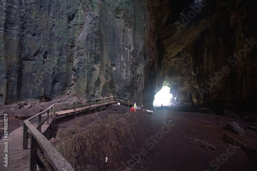 A person exploring a spacious cave with a wooden walkway leading towards the illuminated cave entrance. photo