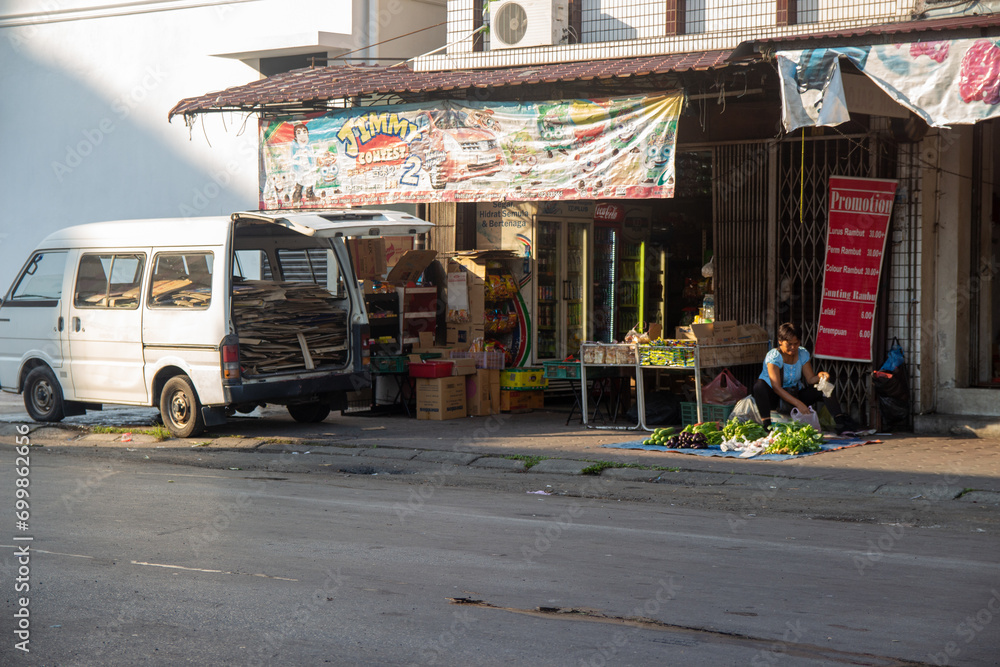 A street vendor sitting by a display of vegetables next to a parked van in front of a small shop