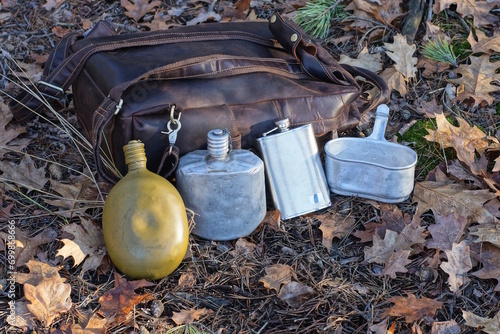 a row of three old flasks and an aluminum pot stand on the ground next to a brown backpack on the street