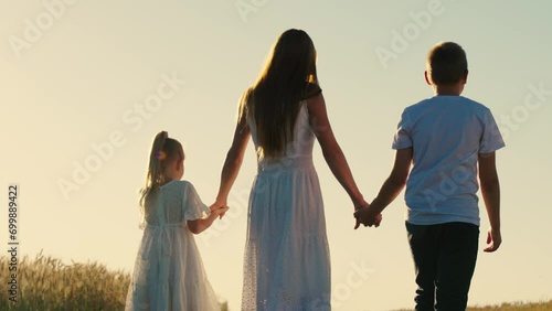 Cheerful mother with children walks among golden wheat fields. Little kids talk with mother enjoying time in fresh air. Happy family members walk holding hands on warm summer day in countryside photo