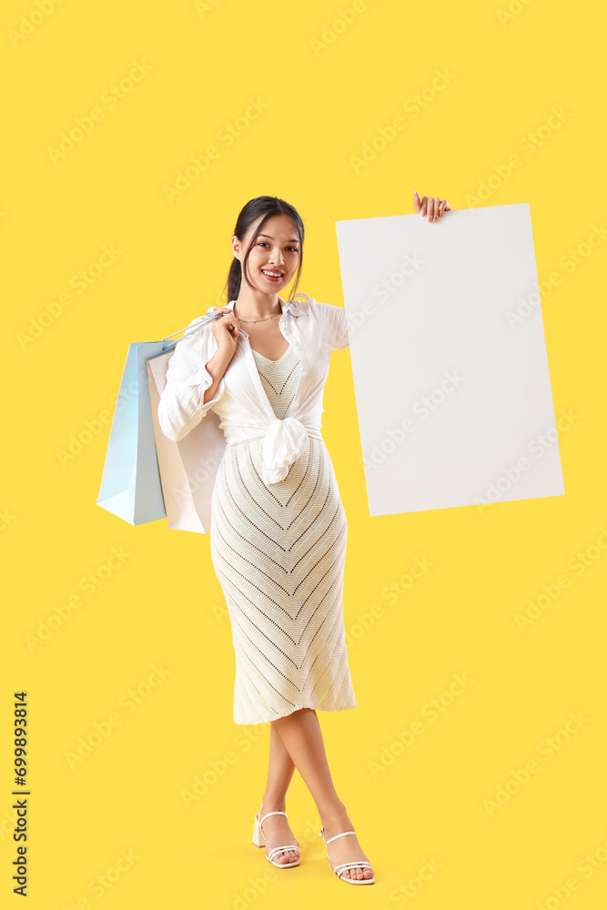 Young Asian seller with blank poster and shopping bags on yellow background Young Asian seller with blank poster and shopping bags on yellow background