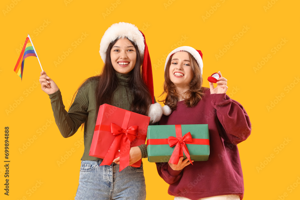 Fototapeta premium Young lesbian couple with Christmas gifts, engagement ring and LGBT flag on yellow background