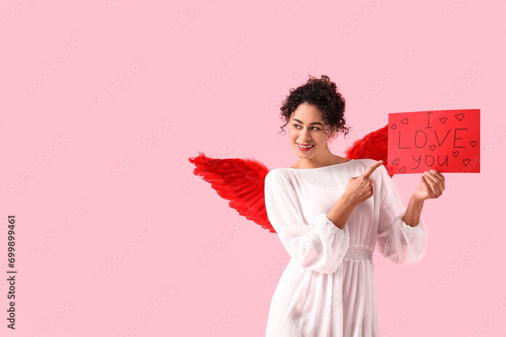 Young African-American woman dressed as Cupid holding paper with text I LOVE YOU on pink background. Valentine's Day celebration Young African-American woman dressed as Cupid holding paper with text I LOVE YOU on pink background. Valentine's Day celebration