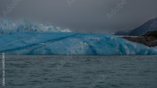 A fragment of a glacier. A wall of blue ice with sharp peaks and cracks and a huge melting iceberg in a glacial turquoise lake. Mountains against a cloudy sky. Fog. Perito Moreno glacier. Argentina. 