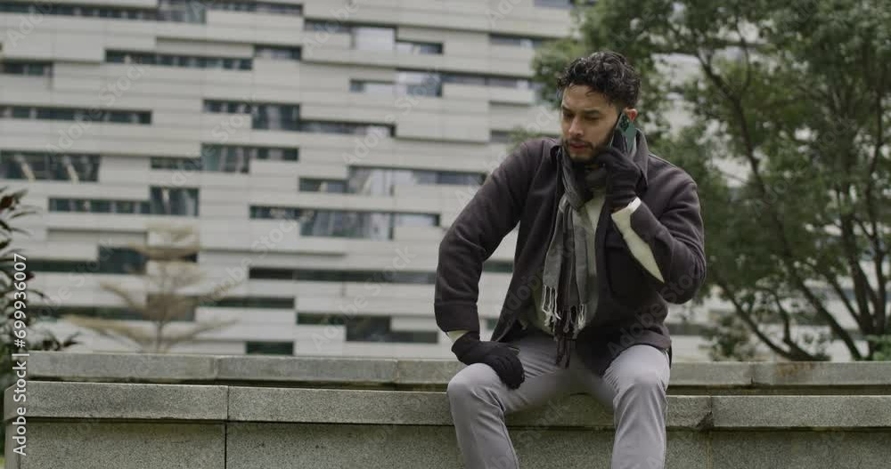 Caucasian male sitting on a bench of a residential district modern area while talking at phone in slow motion. Man in winter clothes outdoors business call Caucasian male sitting on a bench of a residential district modern area while talking at phone in slow motion. Man in winter clothes outdoors business call