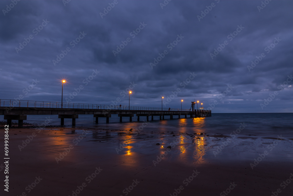 A jetty with lights and reflections in the early morning under a dramatic overcast sky at Mission Beach in tropical Queensland, Australia