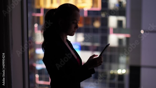 Lady wearing black suit standing at dark room against window, texting using smartphone. Silhouetted half-length shot, blurred office building with bright windows seen blurred on background photo