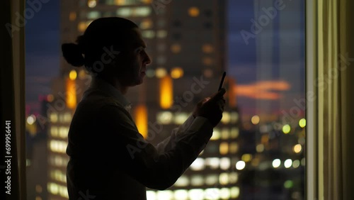 Silhouette of man standing against window, he busy with smartphone. Half length silhouetted shot from side, blurred evening city on background. Man with hipster hair wear shirt photo