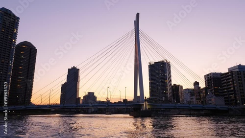 Chuo Ohashi Bridge silhouetted against sunset sky, wide shot from sailing boat. Tall towers and other buildings on banks of Sumida river, characteristic view of Tokyo city from water bus photo