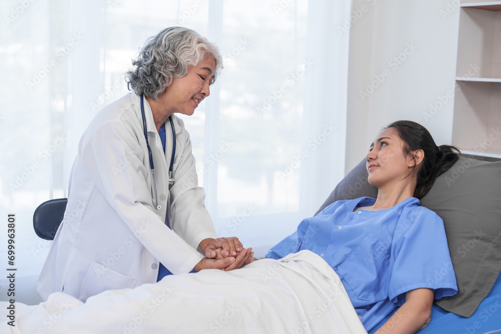 An elderly Asian doctor is smiling and holding the hand of a younger Asian patient who is lying in a hospital bed. An elderly Asian doctor is smiling and holding the hand of a younger Asian patient who is lying in a hospital bed.