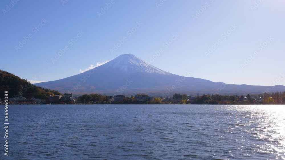 View of Mountain Fuji across Kawaguchiko waters, shot in sunny autumn evening. Majestic mount with snow-capped peak, dark shore and town buildings seen on other side of lake.