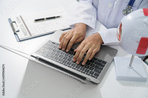 A doctor's hands are seen typing on a laptop keyboard with a stethoscope around the neck on white desk.