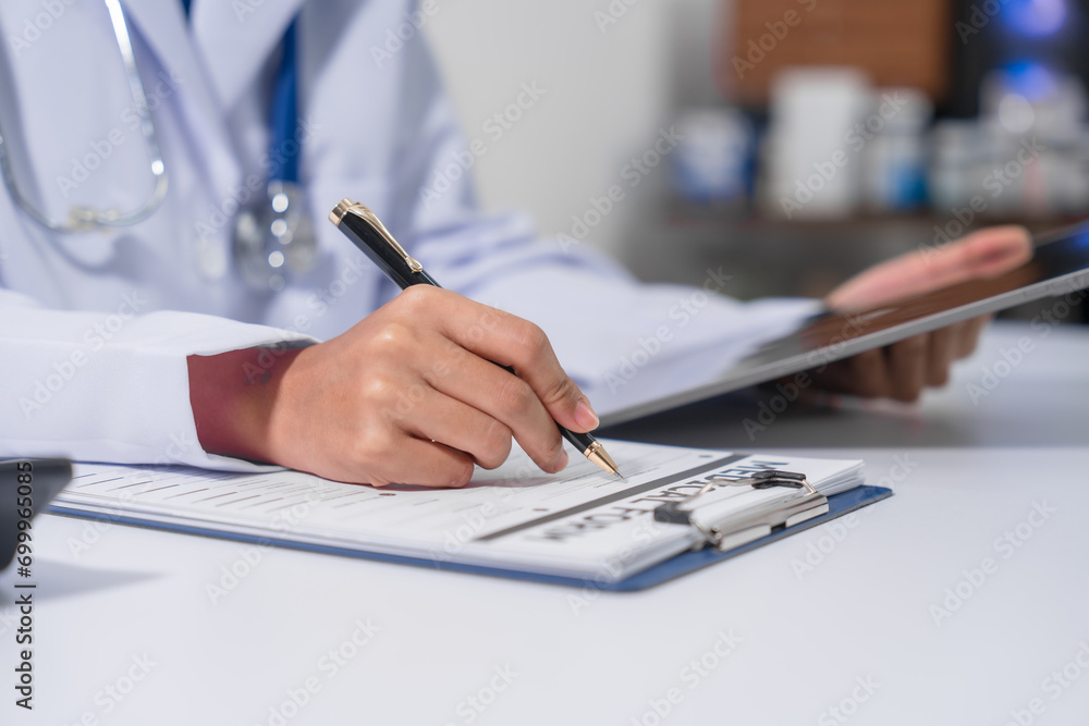 Fototapeta premium A doctor's hand is writing on a medical record form on a clipboard, with a stethoscope and tablet in the background.